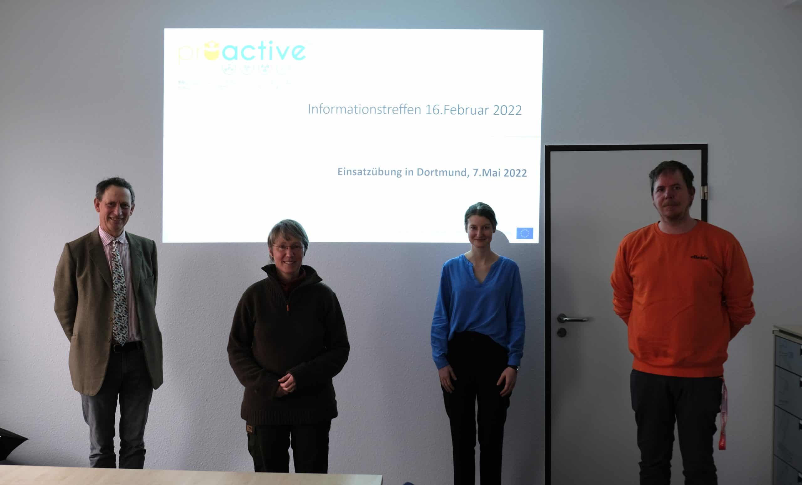 four persons standing socially distanced in a meeting room with the proactive logo projected behind them