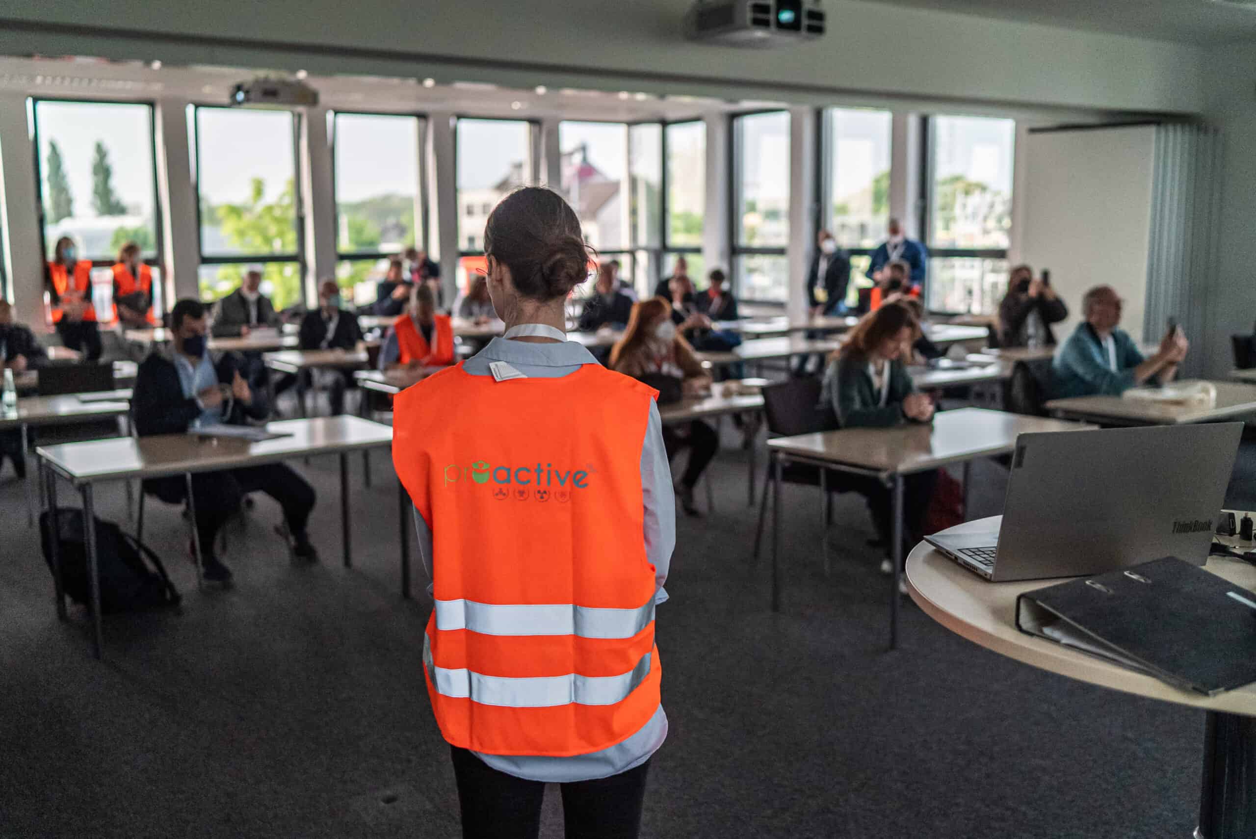 A female PROACTIVE partner wearing an orange waistcoat is standing and looking to participants. In the background, blurred, around 20 people are seated at desks.re sitting