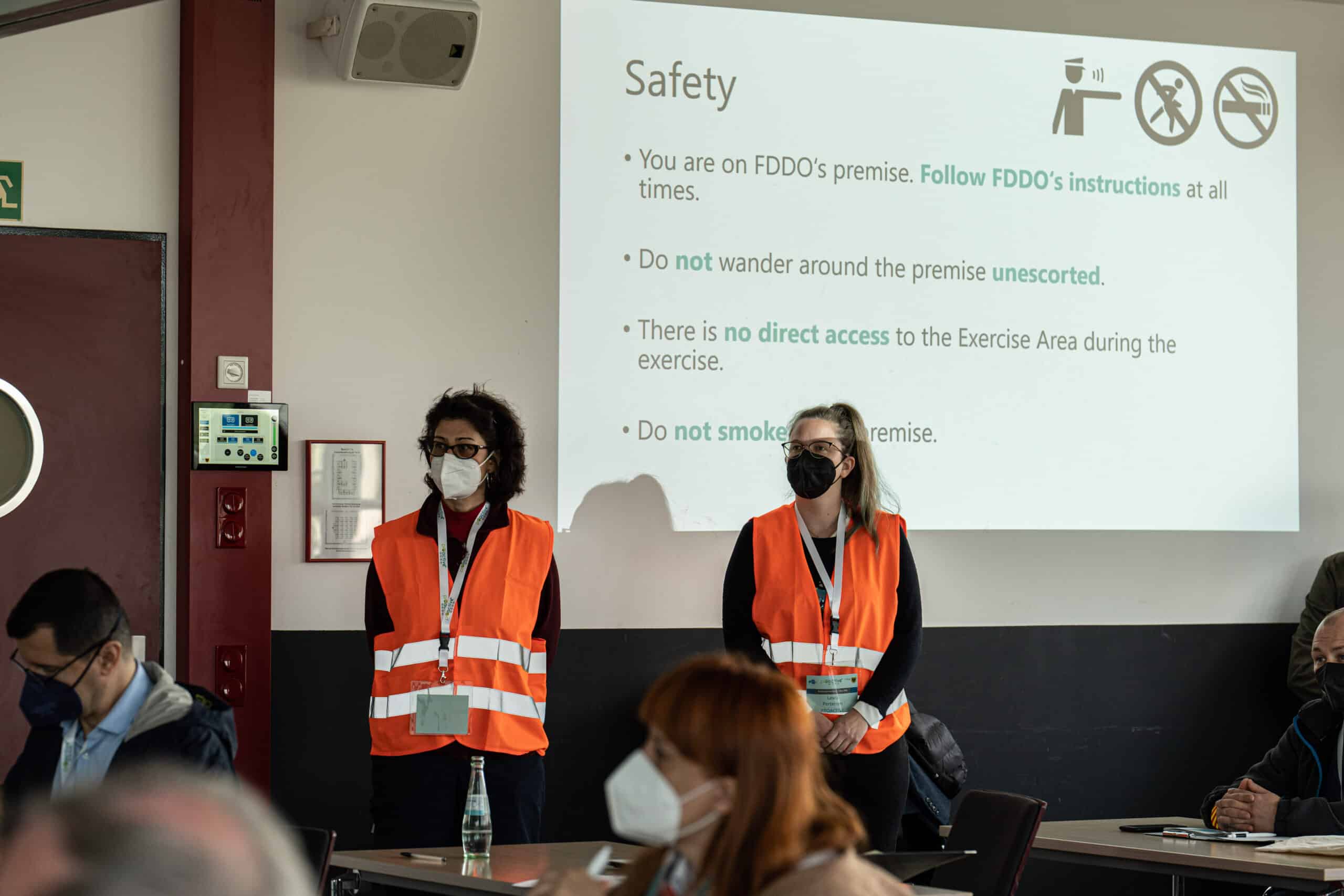 Two female PROACTIVE staff wearing a orange waistcoat are standing in front of the screen where safety instructions are written.