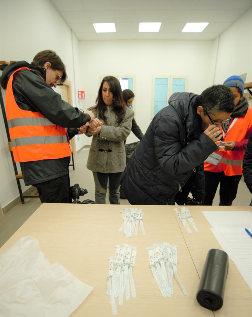 In the foreground a desk with many wrist bands and forms. In the middle ground, two people (a man and a woman) are wearing orange tabards. One is putting a wristband on the wrist of a woman participant. A man participant is leaning over on the desk and reading a form. 
