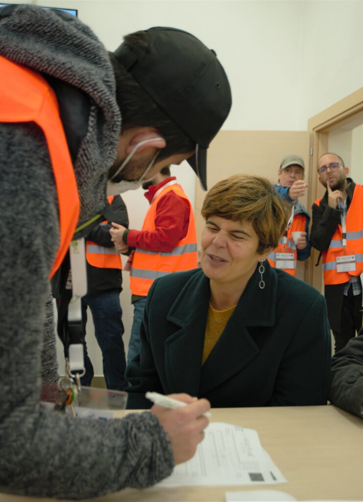 In front a visually impaired woman volunteer is sitting and a person in orange tabard is helping her to fill in the form. In the background many people with orange tabards can be seen in discussion.