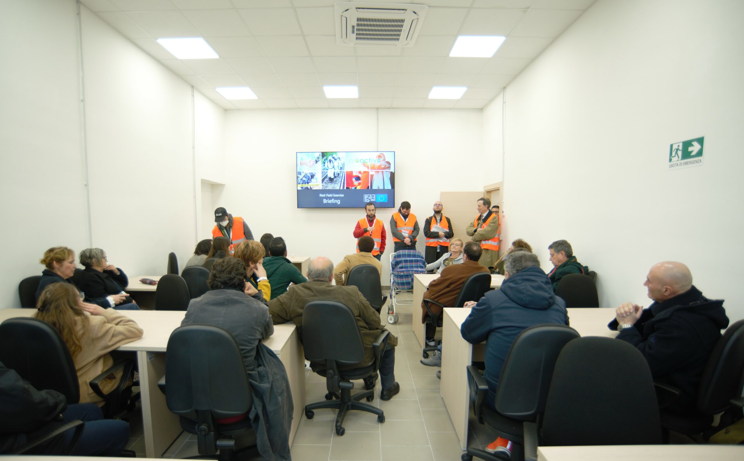 Around 17 people are sitting and listening to people who are wearing orange tabards who are standing in front of participants and one person is explaining something. They are in a classroom. A TV with a non-legible slide is displayed.