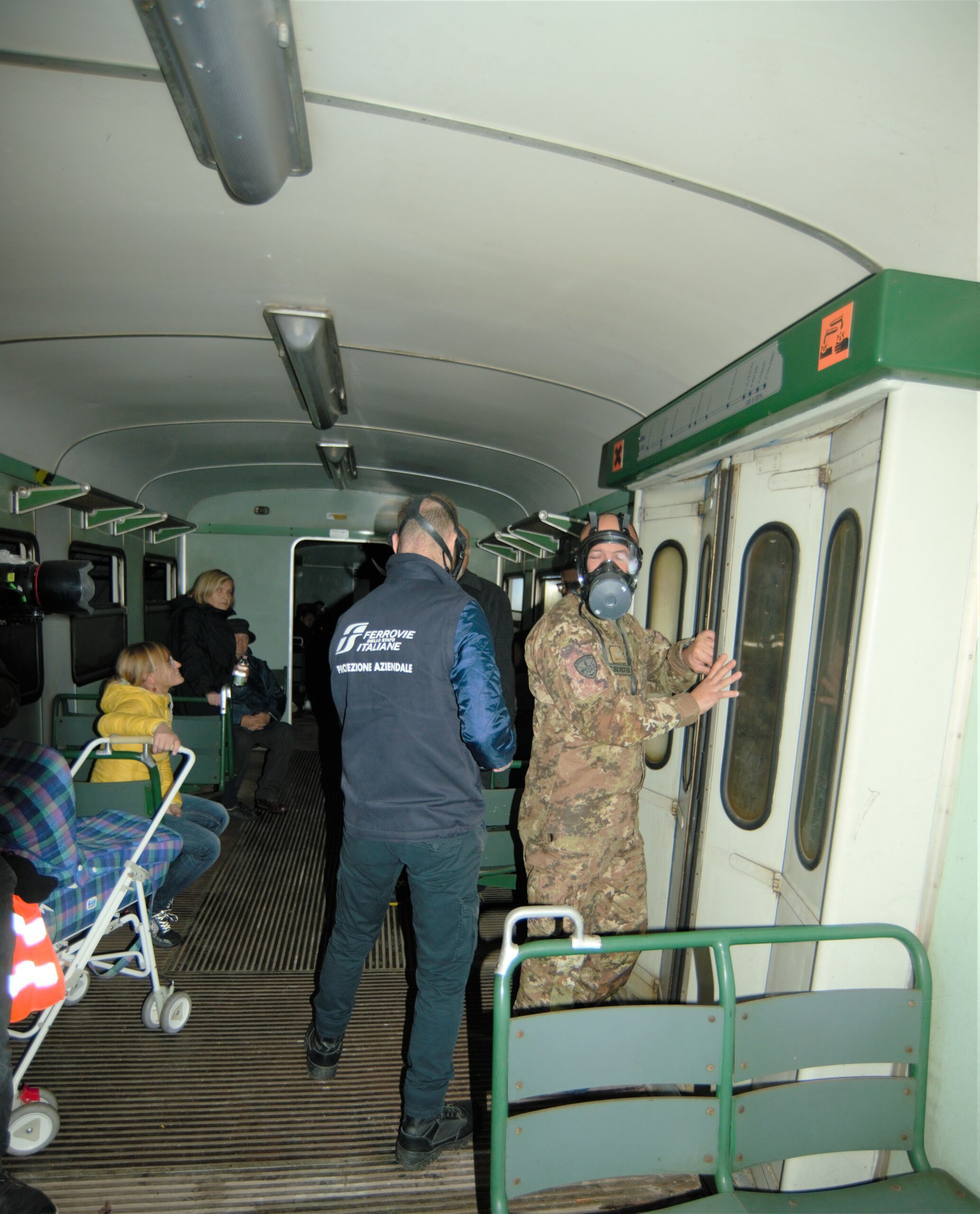 Around 4 people are sitting on train and looking at first responders. Two men wearing respirators are standing close to the train door and a man in an army uniform is trying to open the train door.