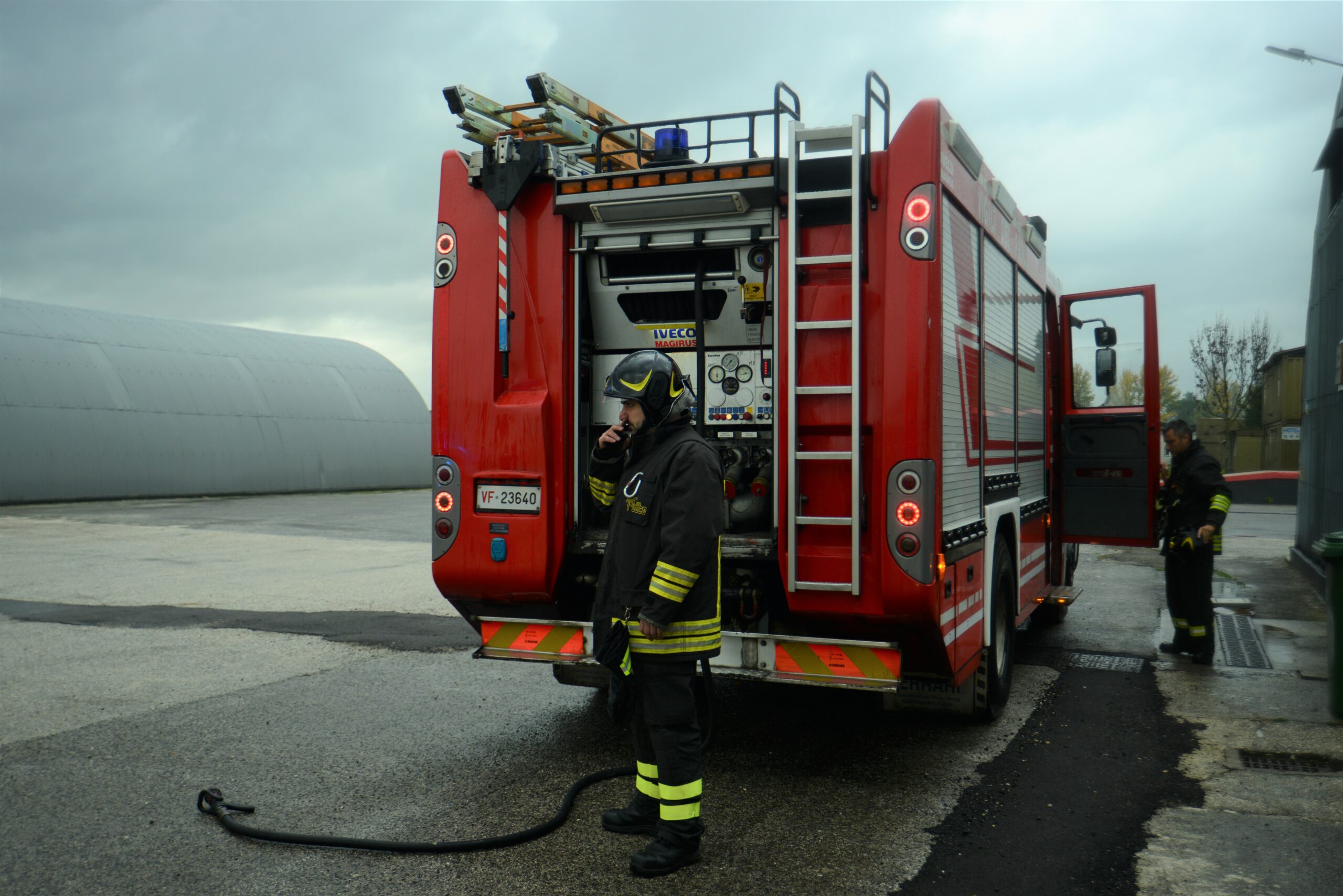 Two firefighters (men) wearing their uniform. One is standing in front and is talking on a radio set. Another firefighter is closing the door of the firetruck.