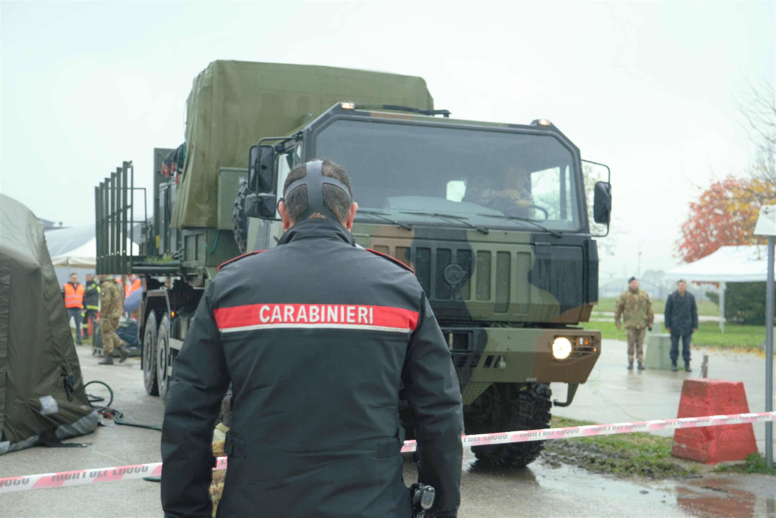 In front of the photo, a man is standing and we see him from behind, on his uniform Carabinieri is written. You can also see black straps on his head which implies he is wearing a mask. In the middle of the photo an army truck/lorry is shown. A man is sitting inside of the truck at the driver's spot.