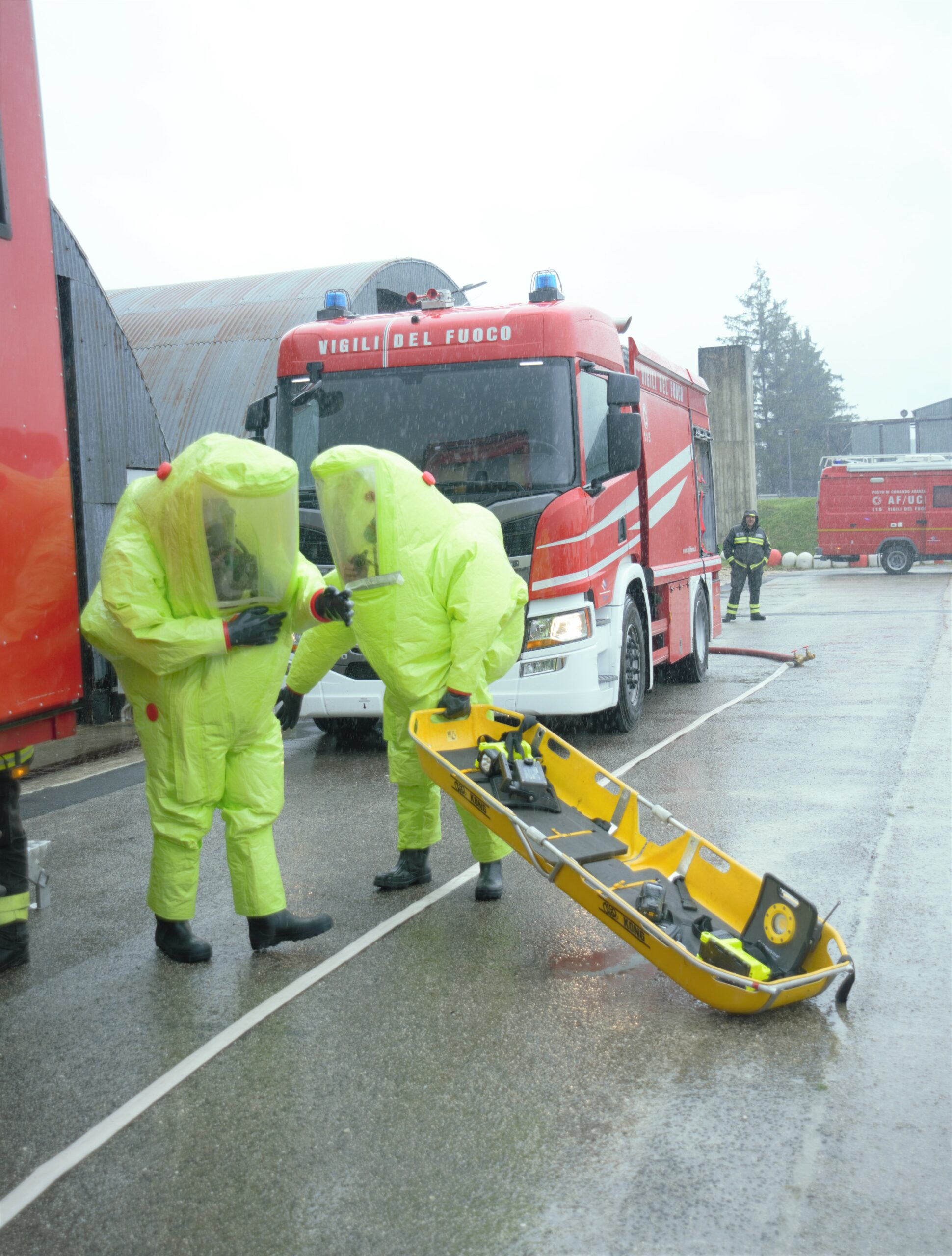 Two persons in a hazmat suit discussing between each other. One of them is holding a yellow bench. Fire trucks can be seen in the background.