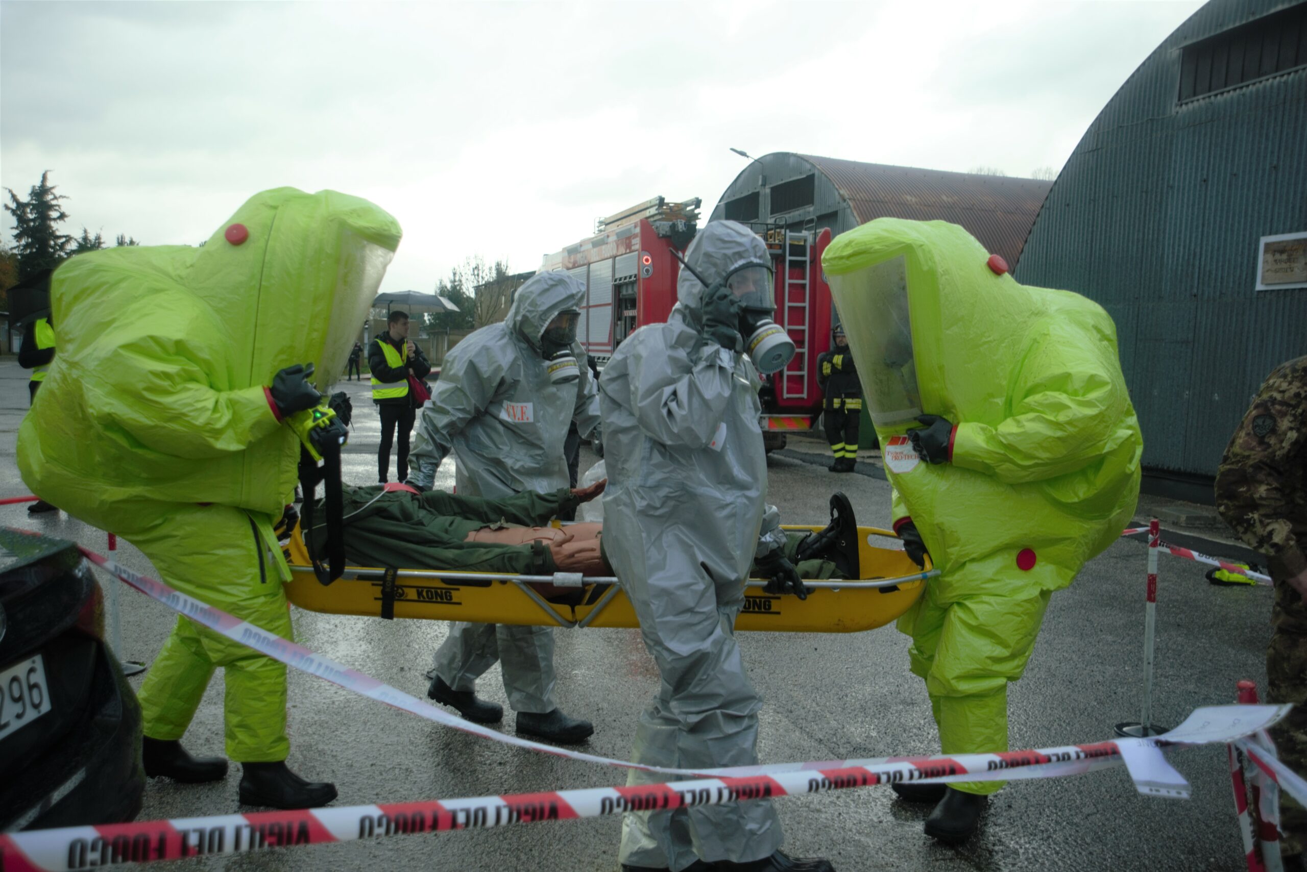 Two persons in a green hazmat uniform and other two are wearing a grey hazmat uniform are evacuating passenger, in this case a mannequin, on bench from the hot zone. The photo is taken outside the platform.