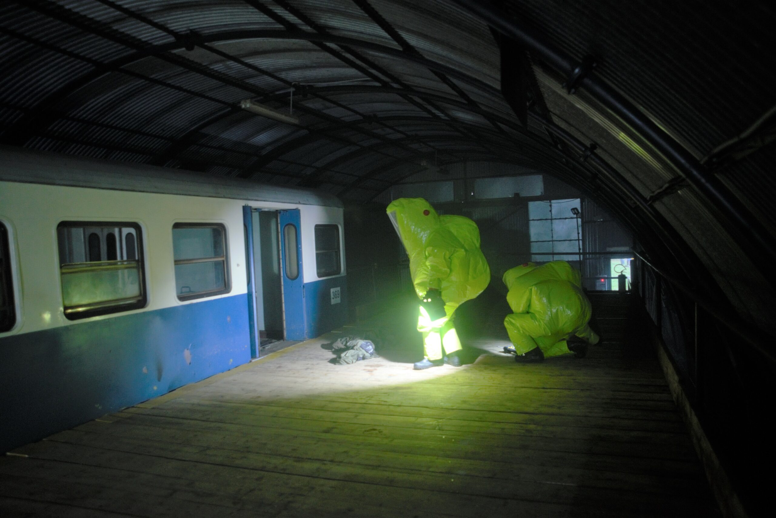 Two CBRN specialised staff wearing a hazmat suit in incident area, on the train platform. One is sitting on one leg and looking to ground with flash at hand. Another is looking to the inside of train. There is something on the ground. The train wagon is visible. 