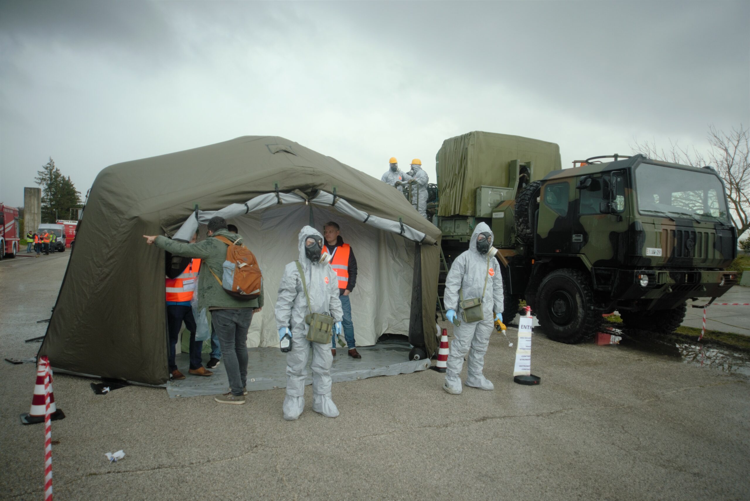 Two persons in a a grey hazmat uniform are standing and behind them there four people inside of tent. Two of them are wearing orange tabards.