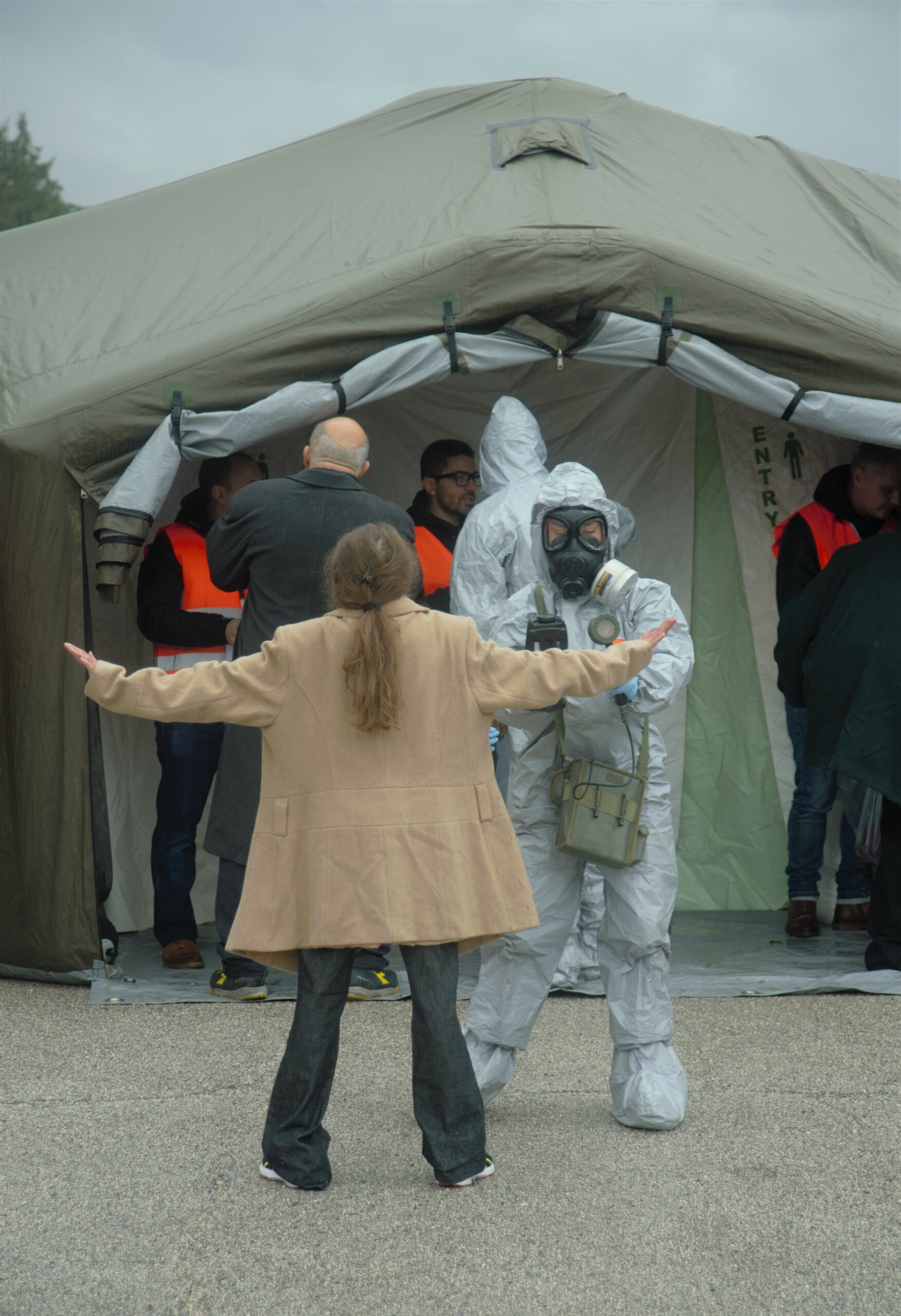 A person in a gray hazmat suit with a detector on his hand is checking hands of a woman participant, who is standing like a starfish (arms and legs spread). Behind them, the decontamination tent can be seen. Another participant is about to walk through the tent, guided by another persons in a grey hazmat suit.