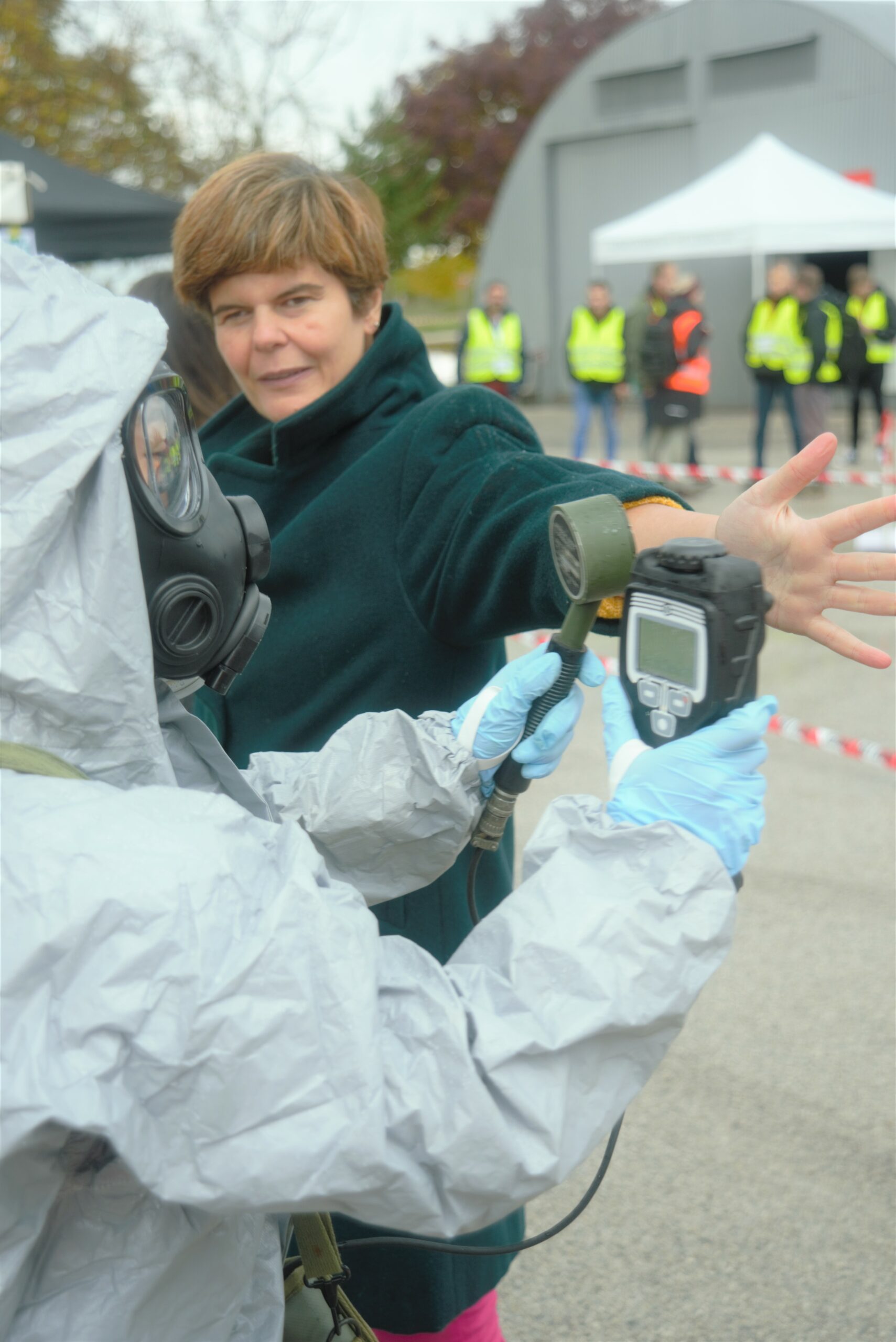 A person in a grey hazmat suit holding a special detector, with the screen in one hand and scanning tool in the other, is holding it up to a woman volunteer's arm. The woman volunteer is looking towards the respondent.