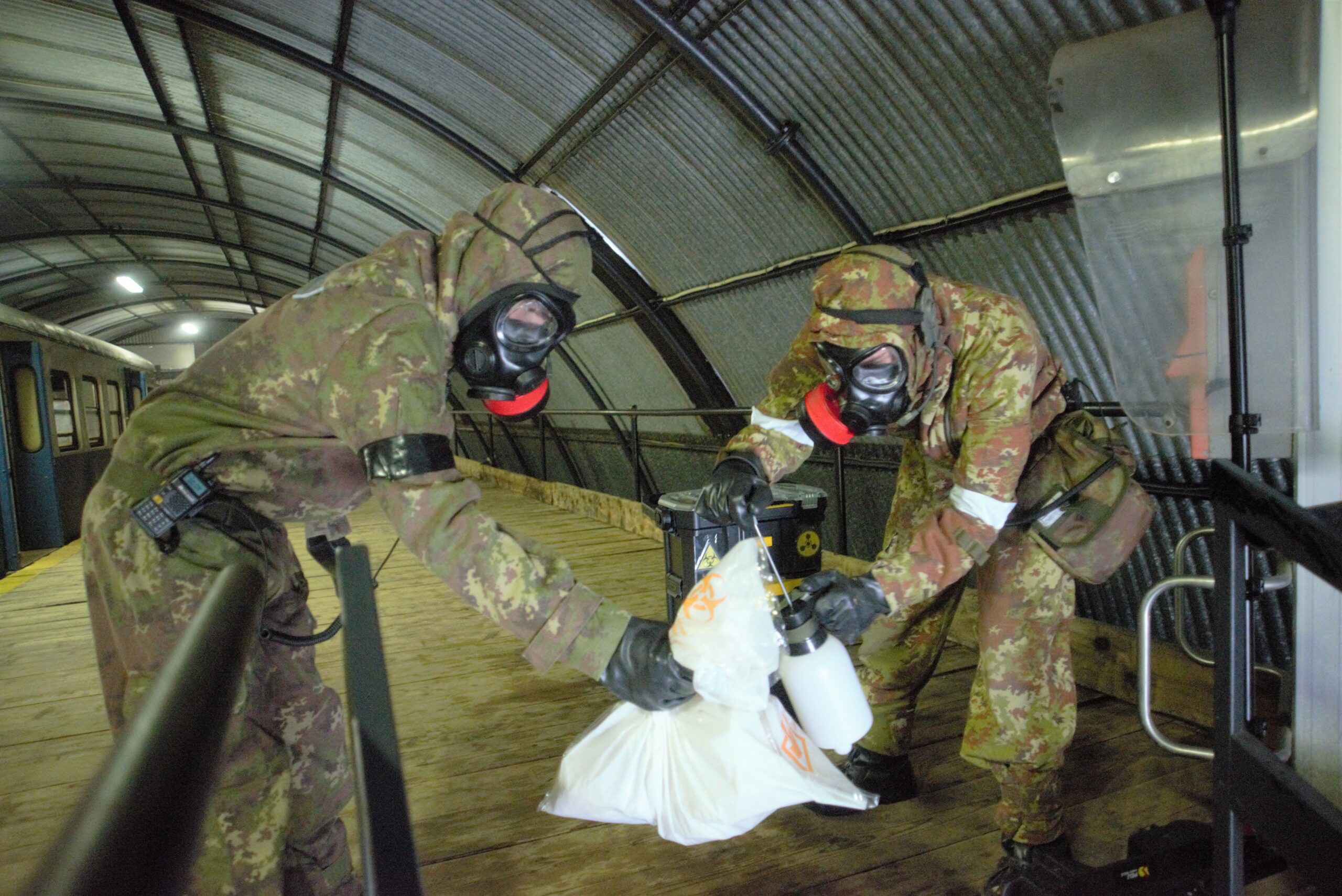 Two persons in a army uniform are wearing a respirator. One of them is holding a white package and another is holding a white bottle .