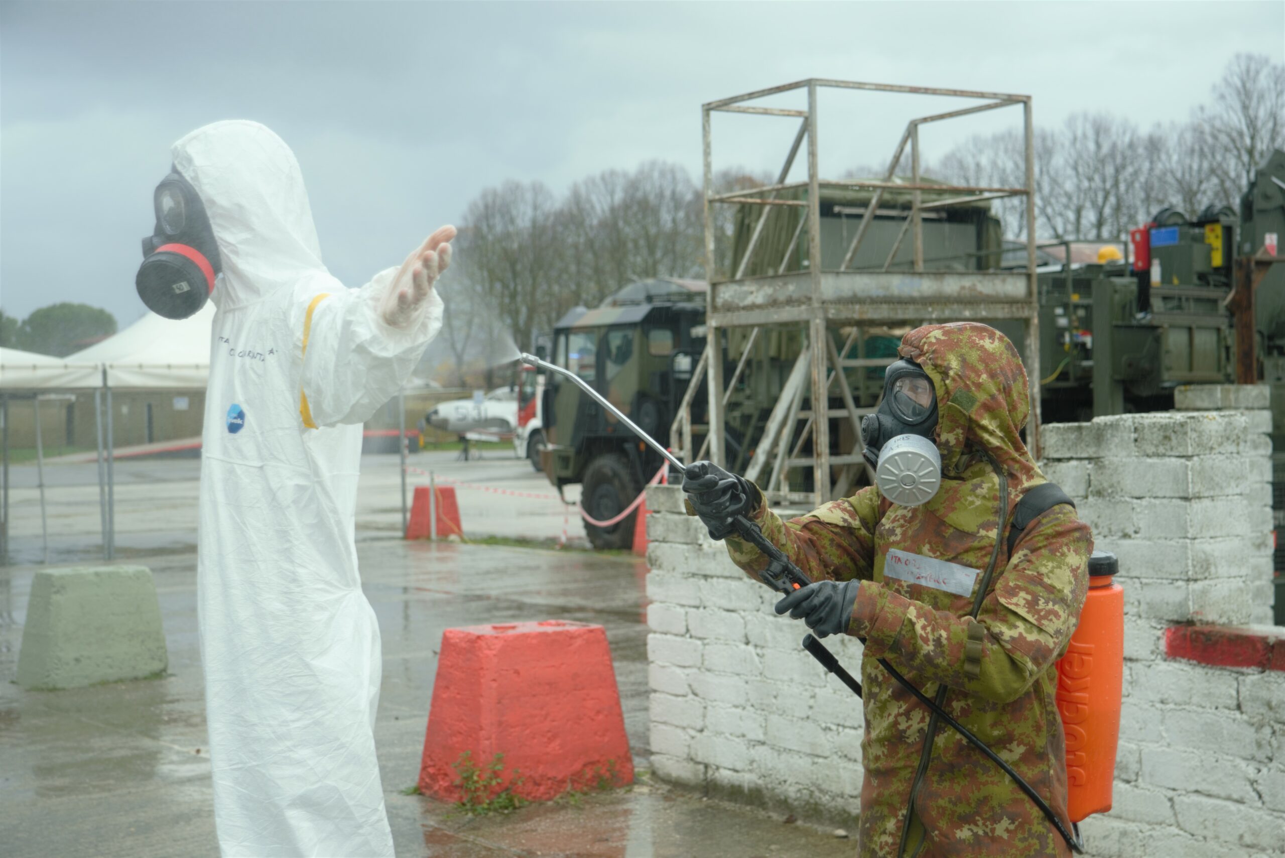 A CBRN Defence Specialist Units from the Army is decontaminating a first responder with water who is wearing a hazmat suit. 
