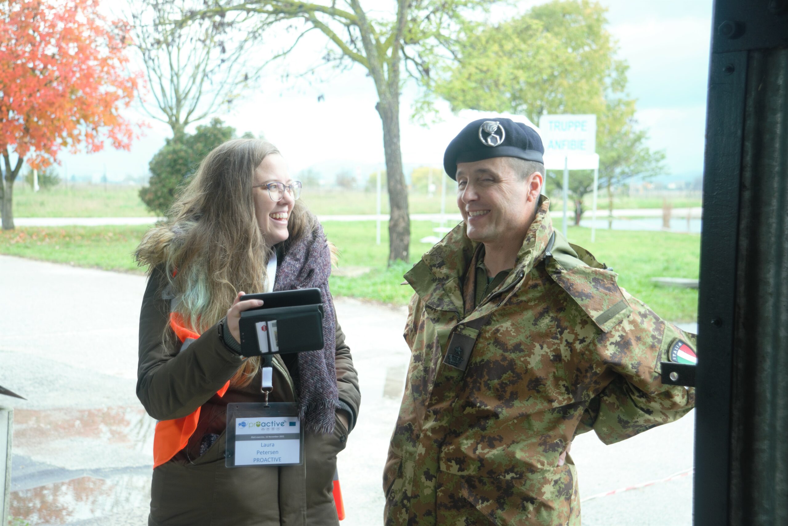Two persons: a woman in orange tabard and man in a military uniform are standing and looking each other and smiling. 