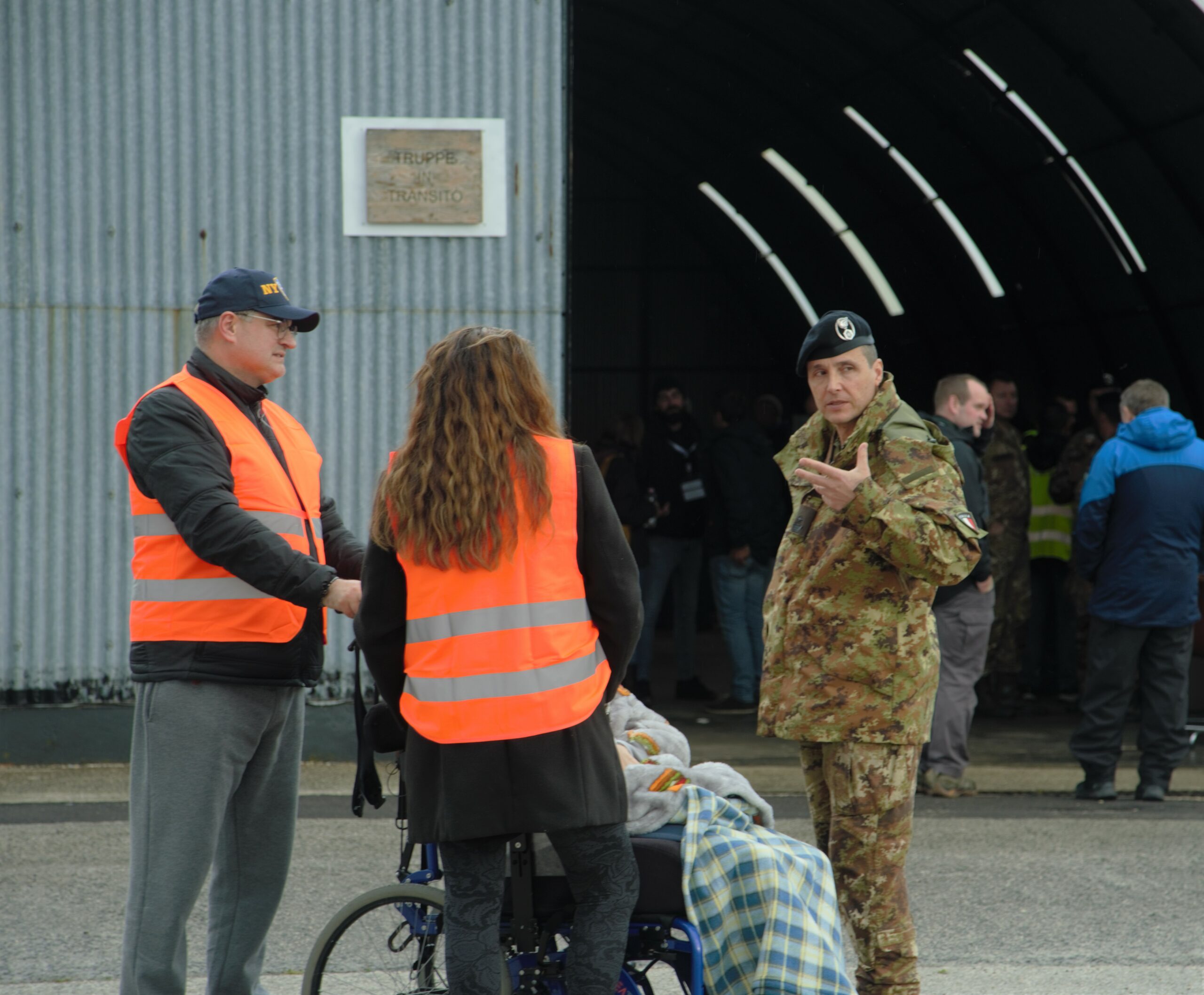 Three people in the front of photo are standing and discussing. One of them is in an army uniform explaining something. A man wearing an orange tabard is holding wheelchair. A woman in an orange tabard is standing with back to the photo.