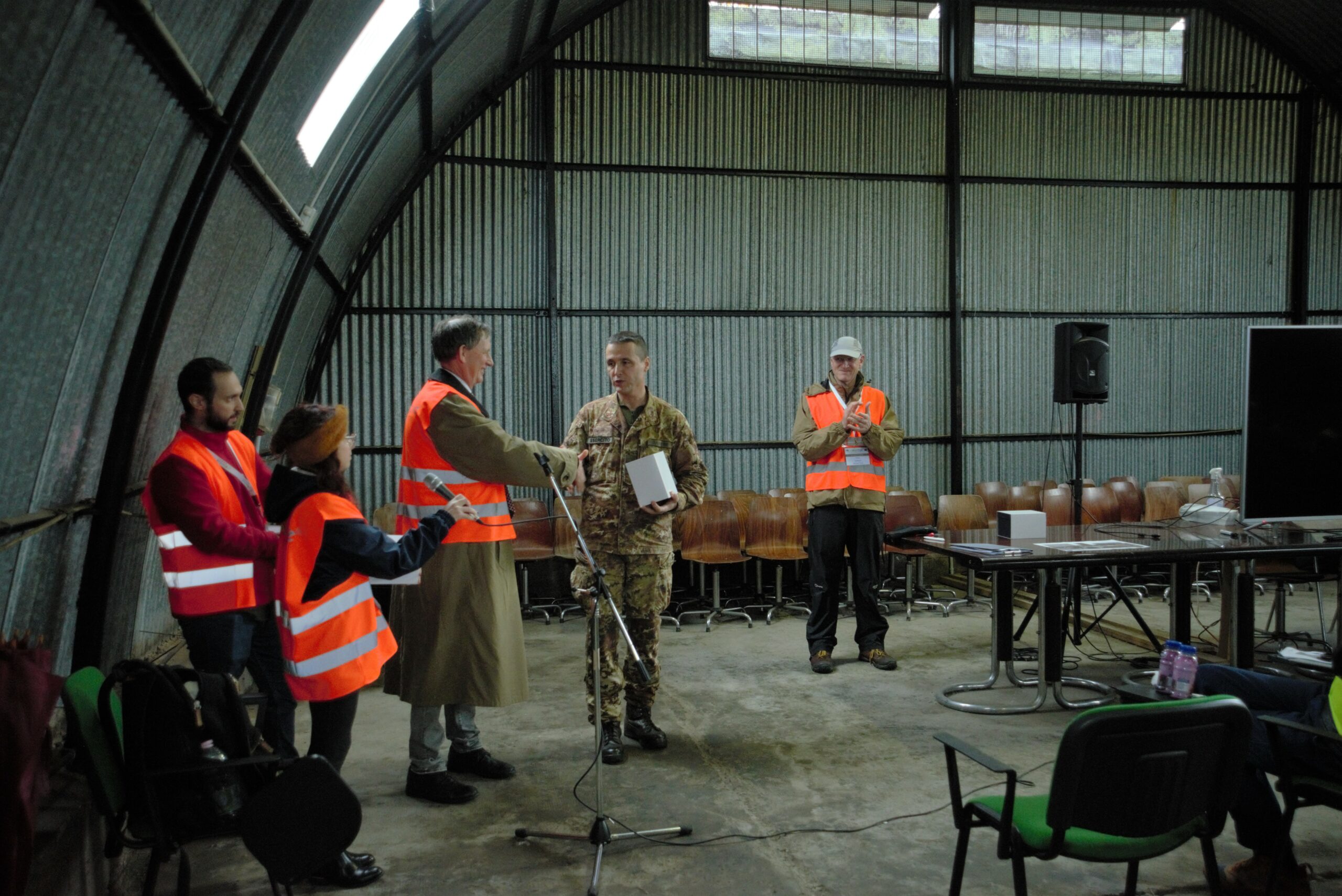 5 people standing in the front of a room. Four of them are wearing orange tabards and one of them is in army uniform. A man in orange tabard in the middle of the photo is shaking hands of a man in army uniform. Two people around them are applausing. A woman in orange tabard is holding a microphone.