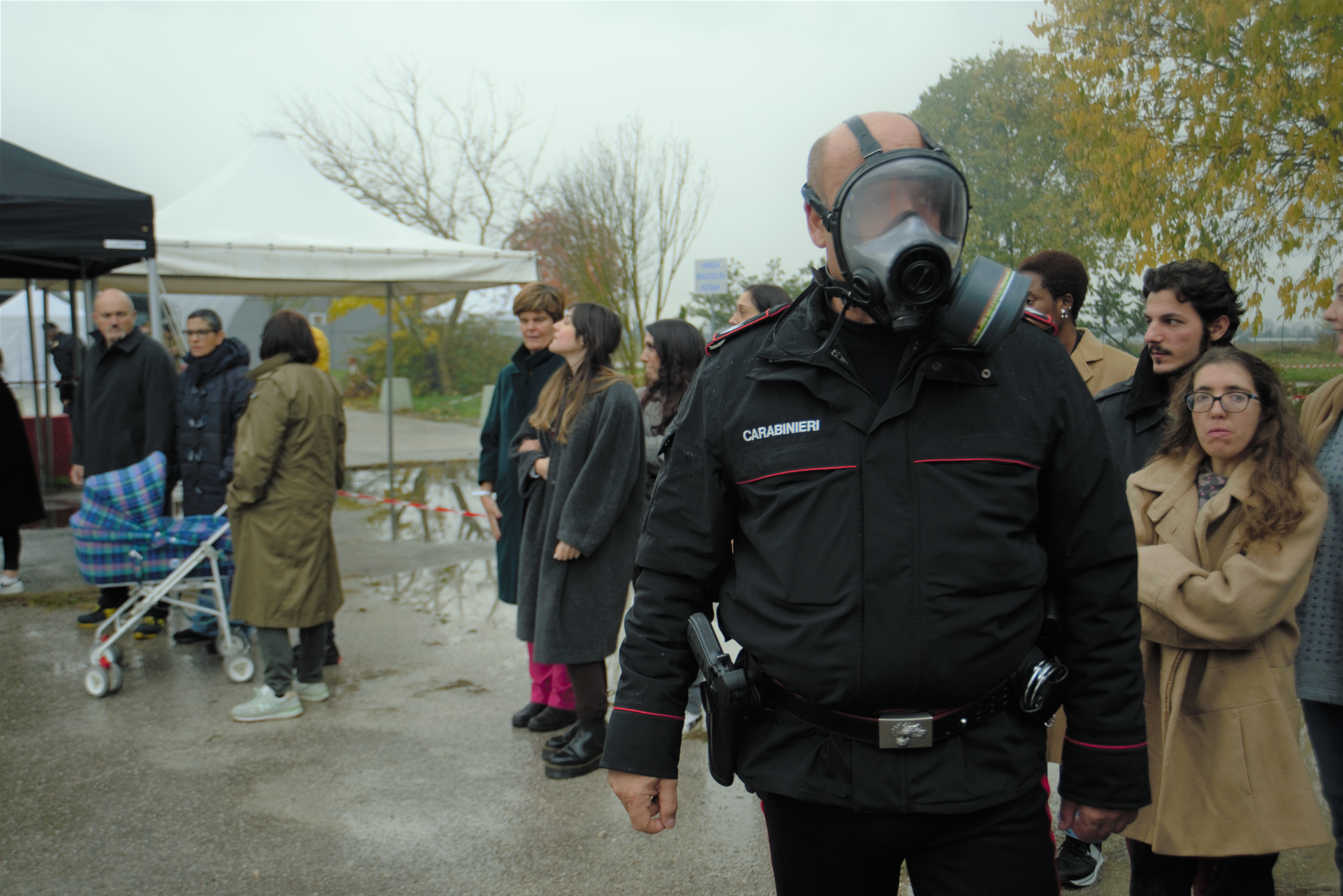 In the foreground, a carabinieri (man) in uniform wearing a respirator. In the background, around 12 volunteer participants are lined up and waiting.. The stroller/pram can be seen.