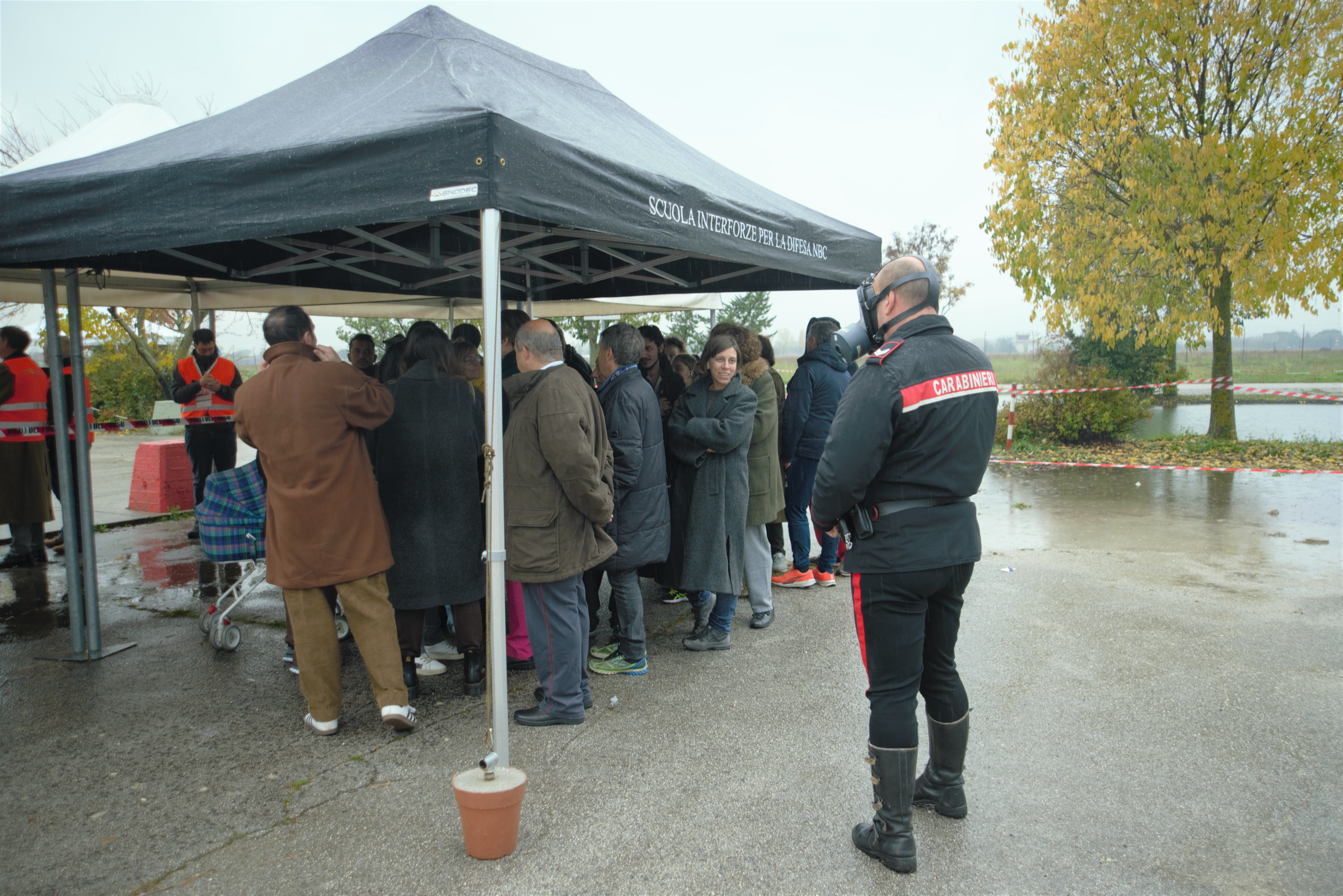 A group of people (around 20) are huddled together under a black tent with no sides. Outside the tent, a carabinieri officer (man) can be seen wearing a respirator. Some people wearing orange tabards are also visible.