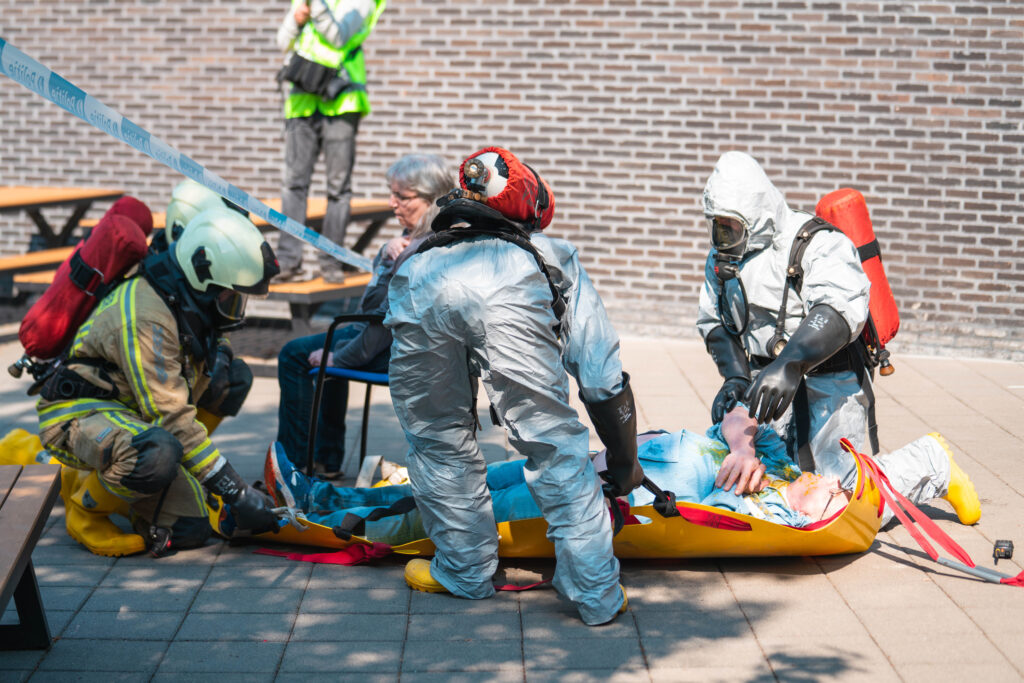 Seven people are in the photo. Two people in grey hazmat suits are going to lift the man lying on the yellow medical bed. Two other people in uniform and wearing helmets are holding a yellow medical bed.