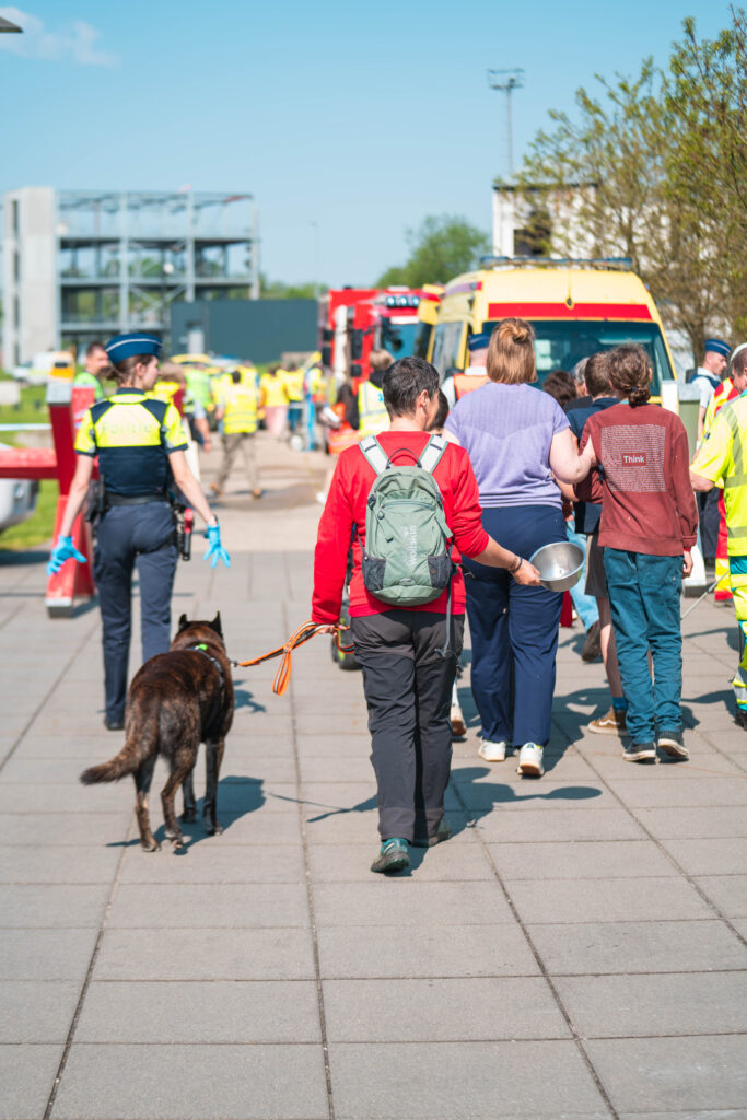 A group of people (different ages) in the photo are walking together. There is also a dog walking next to the woman. Police in uniform are walking next to them and guide them. Caption: Walking toward the medical check