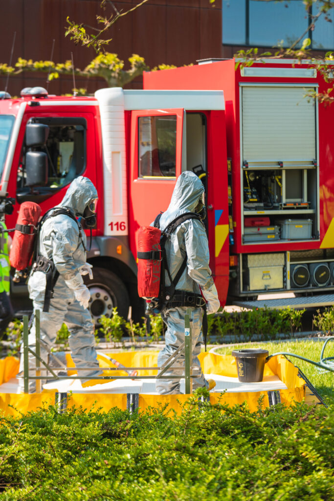 Two people in grey hazmat uniforms are walking outside close to the fire truck.