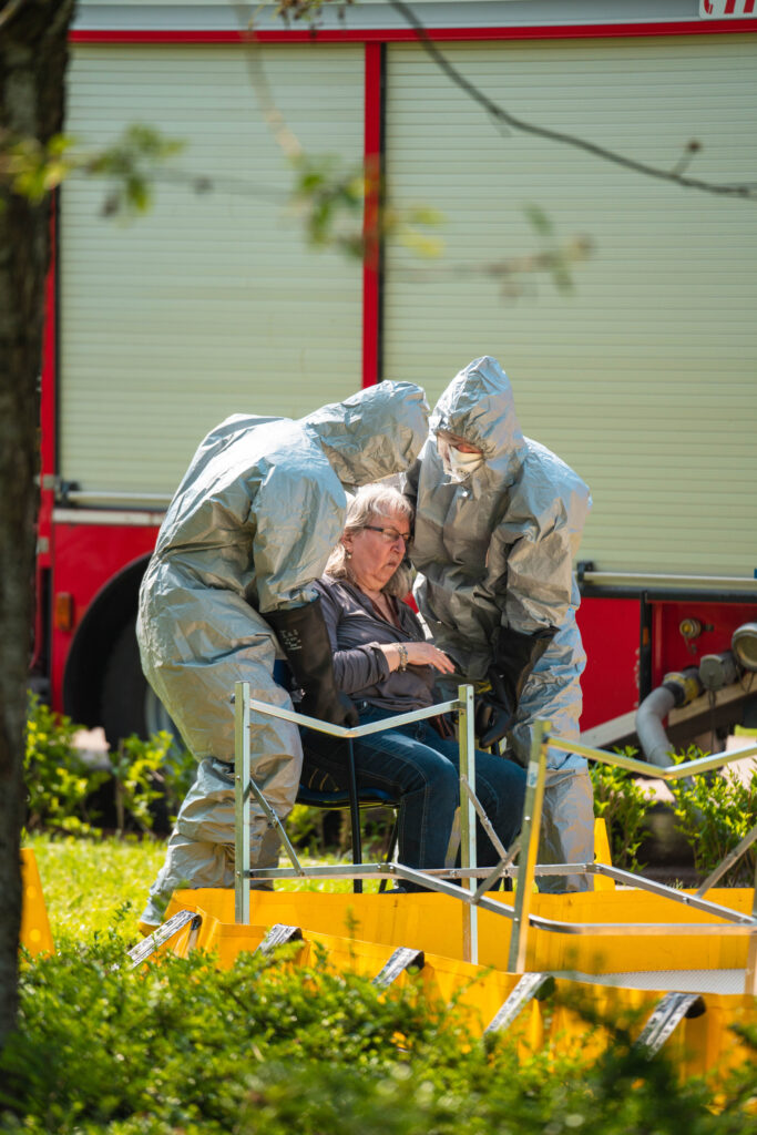 Two people in grey hazmat suits are helping a senior-aged woman sitting on a chair.