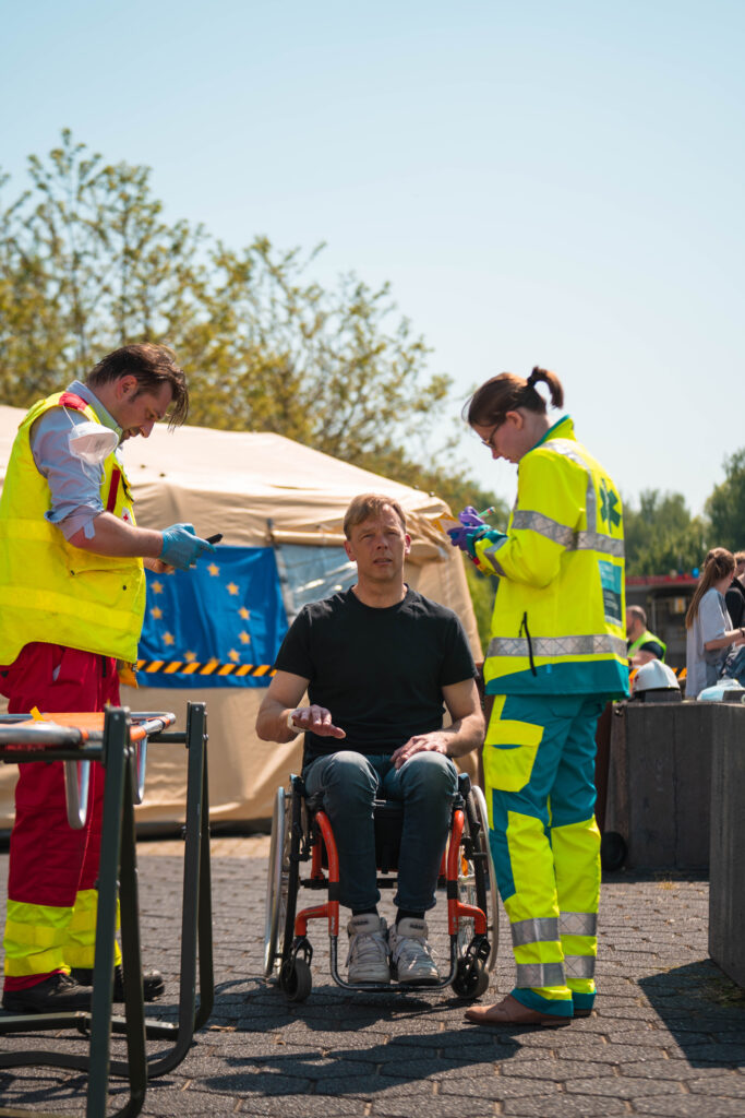 Three people are outside. Two people are in green uniforms filling up something and between them, a man in a wheelchair is explaining something. There is a tent behind them.