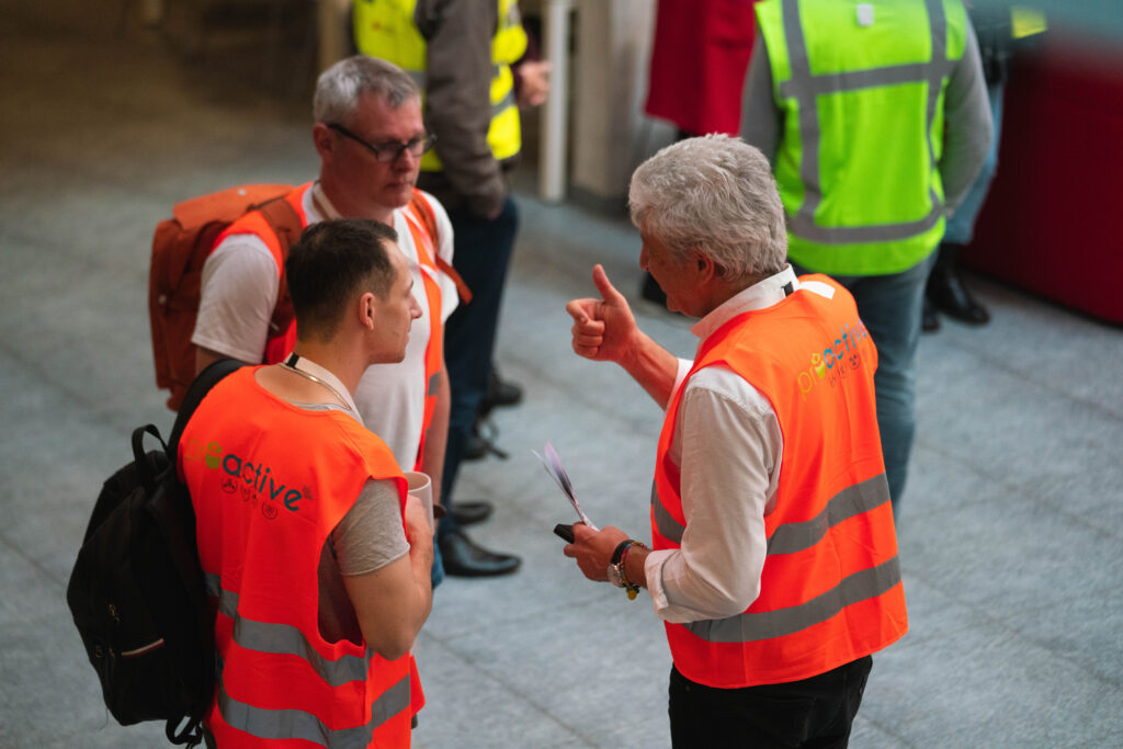 Three men wearing orange waistcoats written PROACTIVE and discussing with each other. Next to them, there are three people in green waistcoats.
