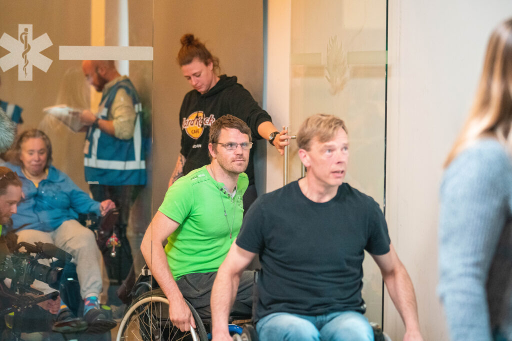 Seven people in the photo (three people: two men and a woman in a wheelchair) Three people are standing. A woman is opening the door and a man wearing a green T-shirt in a wheelchair is entering.