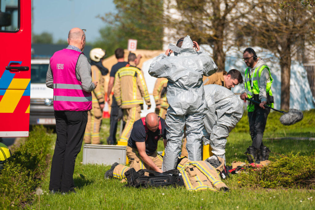 Nine people in the photo. In the front of the photo, a man in a pink waistcoat is standing with his back. A man in a light brown uniform is leaning back and helping to remove a hazmat suit from the man who is standing next to him.