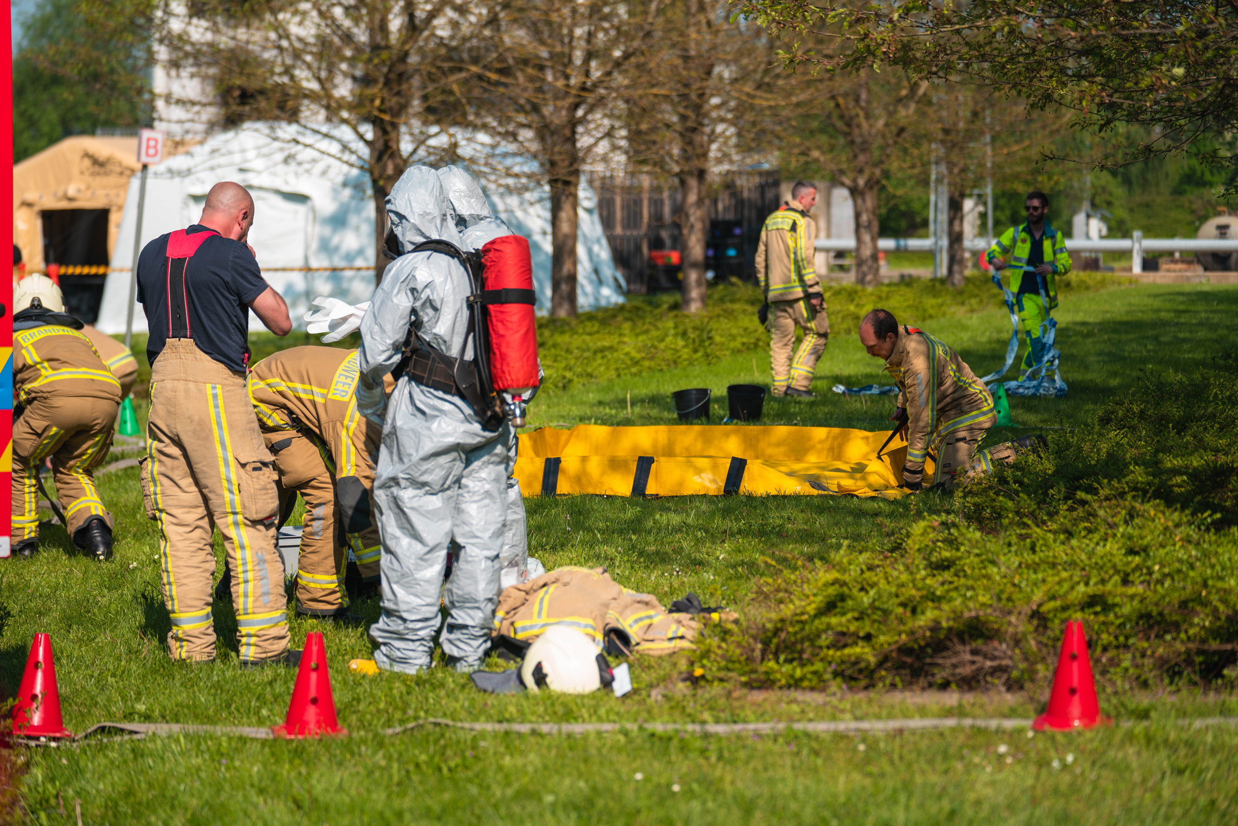 Around nine people in the photo. Six people in light brown uniforms with yellow stripes. Two people in grey hazmat suits. One man in the green waistcoat is holding something.