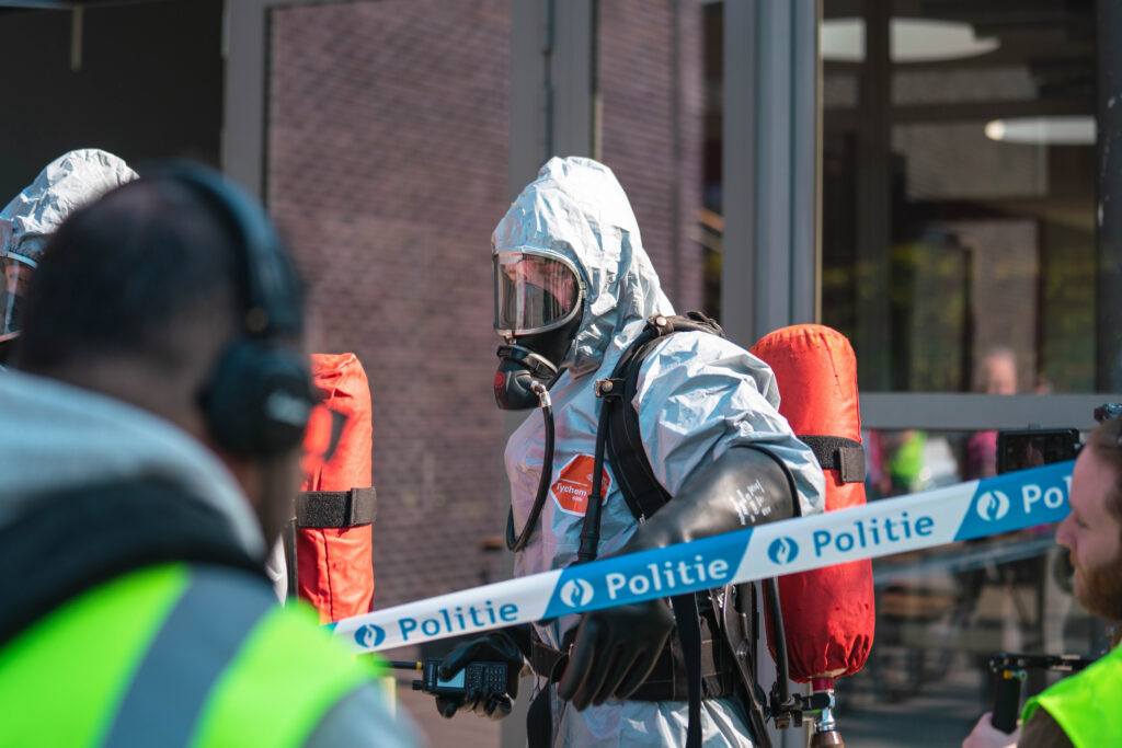Four people in the photo. Two people are in grey hazmat suits. Another two men in green waistcoats are standing opposite them.