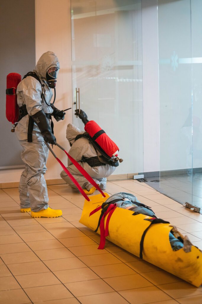 Two people in grey hazmat suits and wearing gear masks. One is holding a red rope which is connected to the yellow roll. Inside the yellow roll, there is a person lying. Another person is leaning down and holding the door with one hand.